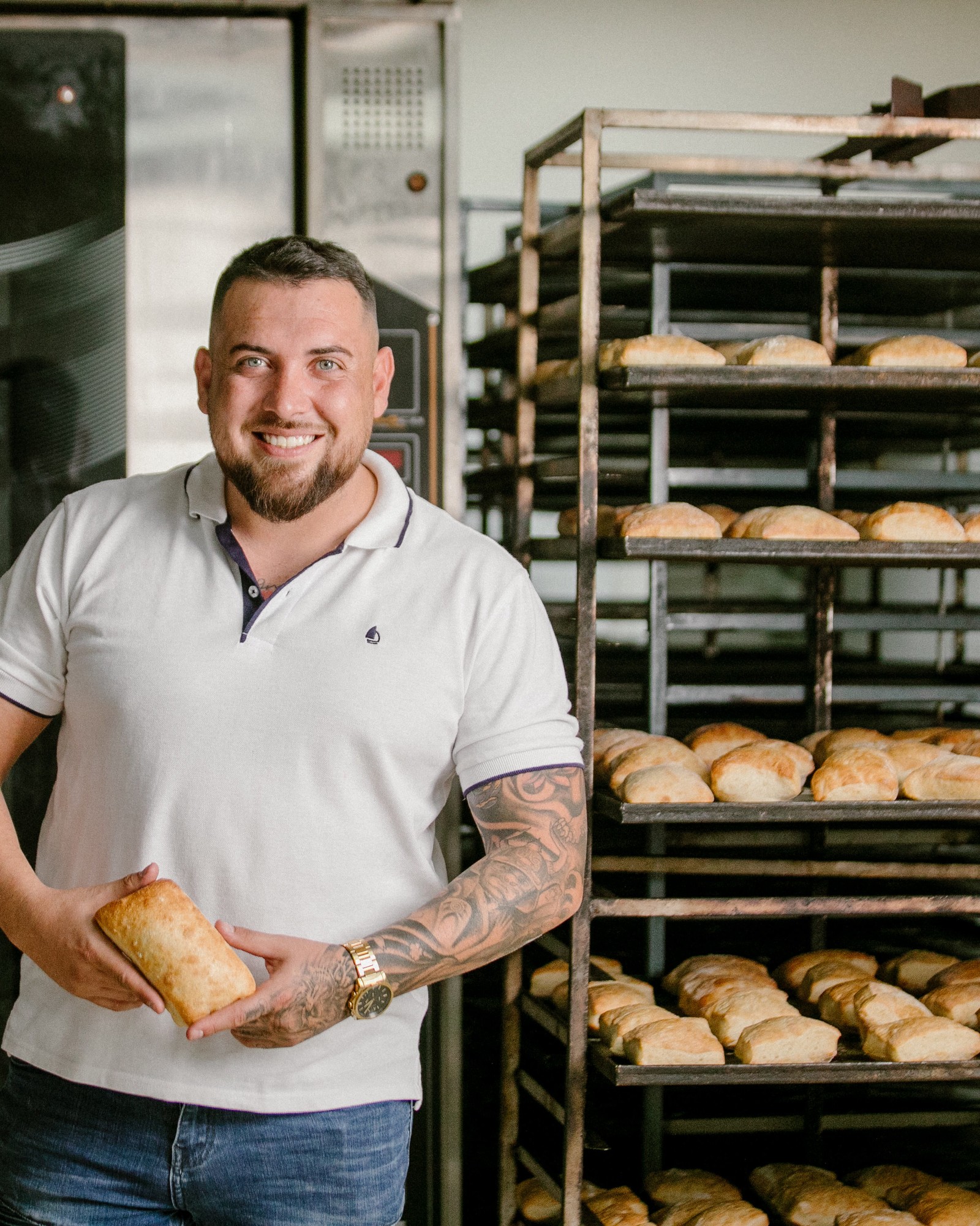 Rodrigo, the owner of El Chapatero, smiles holding a freshly baked chapata beside a metal rack full of bread in his bakery.