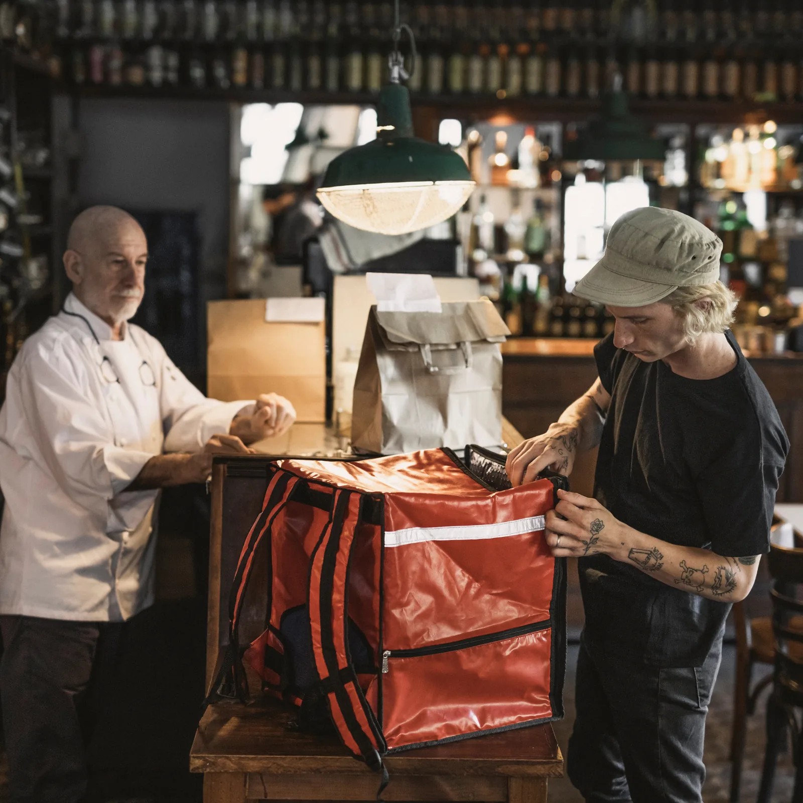 Inside a restaurant, a cook in whites hands a paper bag across the counter while a courier in a cap opens a red insulated box on a wooden stand.