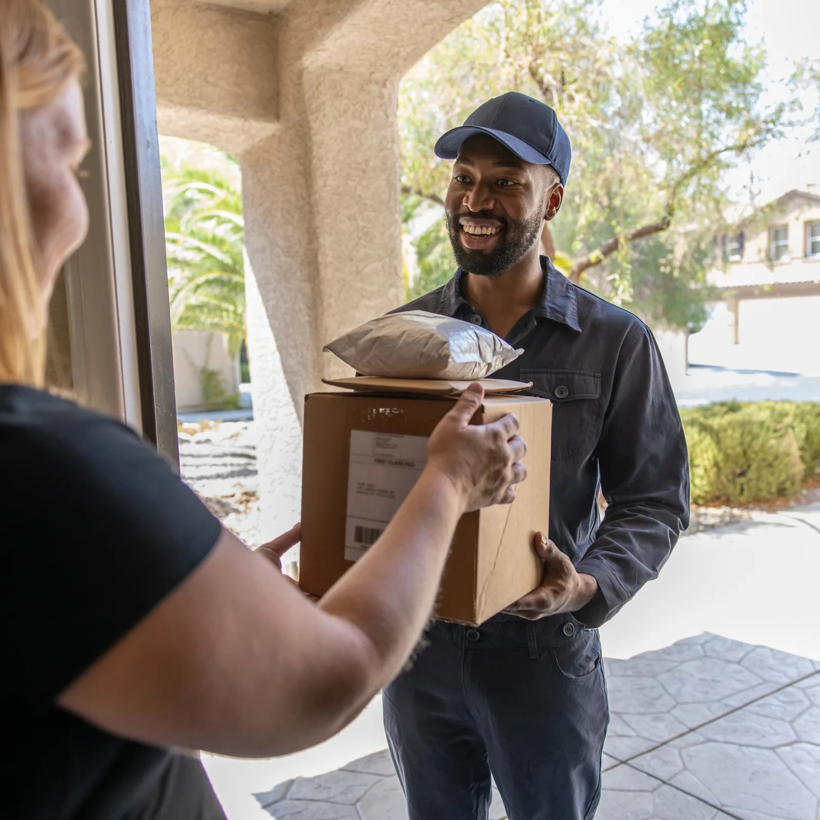 A courier in a cap smiles at a doorway as he hands a cardboard box and a package to the person who opened the door.