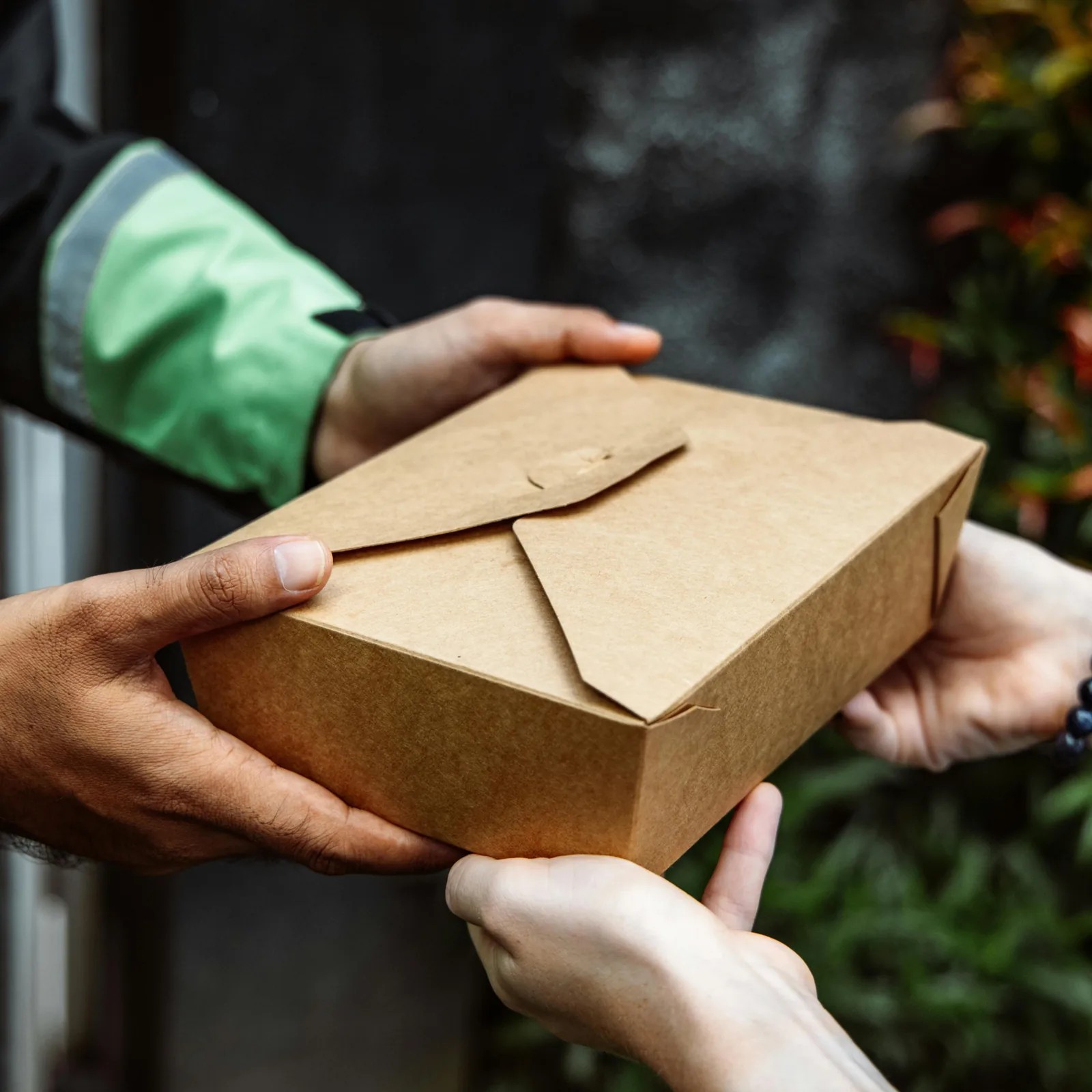 Close-up of a kraft food box passing from a courier's hands into the customer's at a doorway.
